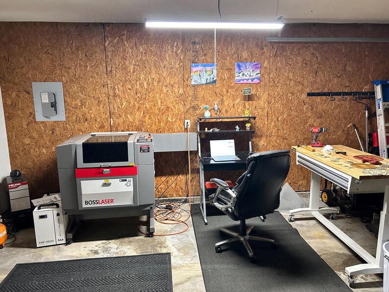 May include: A workshop space featuring a gray Boss Laser machine, a black office chair positioned at a desk with a laptop, and a wooden workbench. The room has wood-paneled walls, a fluorescent overhead light, and various tools and equipment.