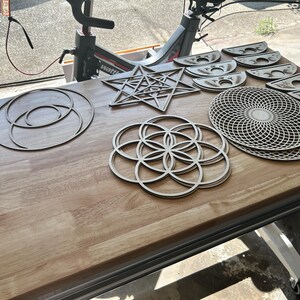 May include: A collection of laser-cut wooden geometric shapes including a large star, a circular flower of life pattern, a complex grid disc, and several crescent moon phases. These light-colored wooden pieces feature intricate cutouts and designs.