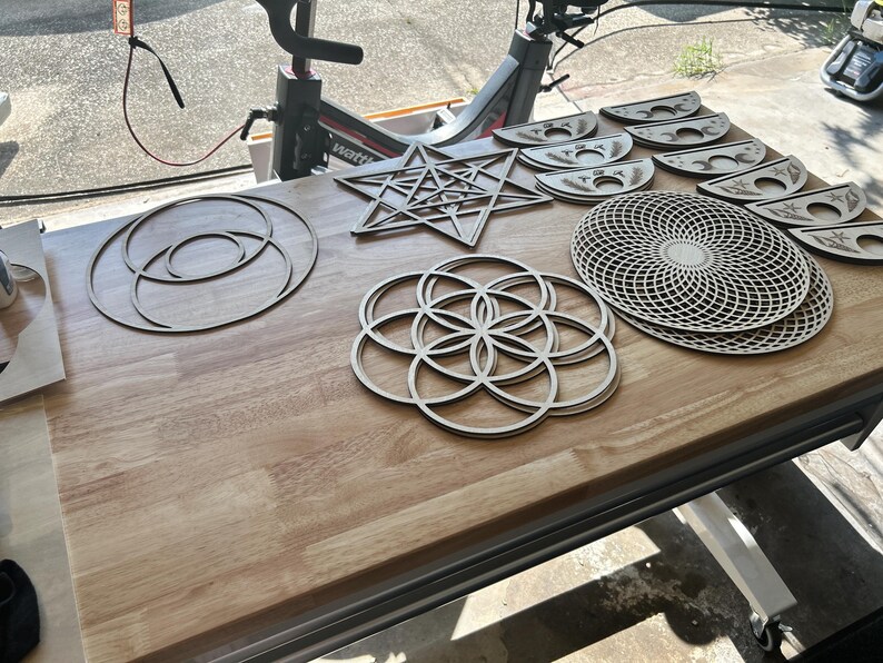 May include: A collection of laser-cut wooden geometric shapes including a large star, a circular flower of life pattern, a complex grid disc, and several crescent moon phases. These light-colored wooden pieces feature intricate cutouts and designs.