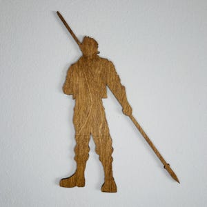 May include: Wooden silhouette of a person holding a spear. The figure is in a standing pose, with the spear angled across the body. The wood has a warm, natural tone, and the figure is against a white background.