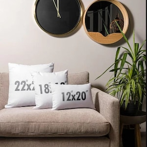 May include: Three white throw pillows with black text on a beige couch. The pillows are labeled 22x18, 18x18, and 12x20. There are two gold and black clocks on the wall behind the couch.