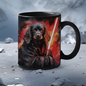 Puede incluir: Taza de cerámica negra con una ilustración de un perro vestido con un disfraz de ciencia ficción sosteniendo un sable de luz. El fondo es rojo y naranja. La taza tiene un asa negra y está sobre un fondo nevado.