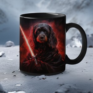 Puede incluir: Taza de cerámica negra con un perro de pelo oscuro sosteniendo una espada láser roja sobre un fondo rojo oscuro y negro. El perro lleva una túnica negra con capucha.