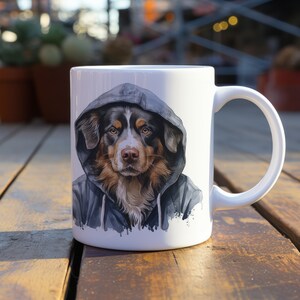 Könnte beinhalten: Weiße Keramiktasse mit einer bunten Illustration eines Hundes, der einen Kapuzenpullover trägt. Der Hund hat braunes, schwarzes und weißes Fell.