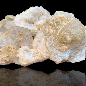 Puede incluir: Un grupo de formaciones minerales translúcidas de color blanquecino con una estructura cristalina. El mineral tiene una apariencia en capas, con algunas áreas que parecen más opacas y otras más transparentes. El fondo es negro, proporcionando contraste.