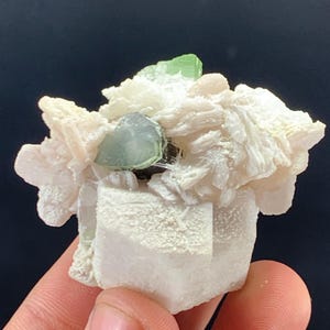 Może przedstawiać: Zbliżenie na skupisko surowych minerałów. Zawiera jasnozielony kryształ, uzupełniony białymi, różowymi i szarymi formacjami mineralnymi. Okaz trzymany jest w dłoni na ciemnym tle, co podkreśla kolory i tekstury.