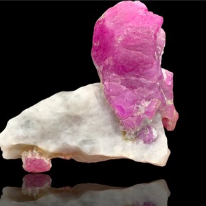 May include: A large, pink ruby crystal sits on a white, rough-hewn rock. The ruby has a deep, rich pink colour and is translucent. The rock is white and has a rough, uneven surface.