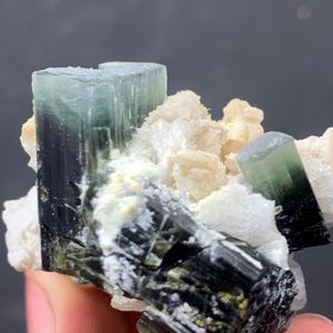 Może przedstawiać: Zbliżenie okazu mineralnego z skupiskiem kryształów turmalinu. Kryształy wykazują gradient kolorów, przechodząc od czarnego do zielonego i kończąc na jasnoniebieskim odcieniu. Białe i beżowe minerały otaczają turmalin.