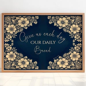 Op de afbeelding: Een ingelijste print met een marineblauwe achtergrond en bloemmotieven in de hoeken. De tekst "Give us each day OUR DAILY Bread" is geschreven in een elegant lettertype. De lijst is lichtbruin.