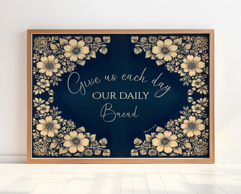 Op de afbeelding: Een ingelijste print met een marineblauwe achtergrond en bloemmotieven in de hoeken. De tekst "Give us each day OUR DAILY Bread" is geschreven in een elegant lettertype. De lijst is lichtbruin.