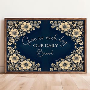 Op de afbeelding: Een ingelijst kunstwerk met een donkerblauwe achtergrond en bloemmotieven in de hoeken. De tekst "Give us each day OUR DAILY Bread" is geschreven in een elegant lettertype. De lijst is donkerbruin.