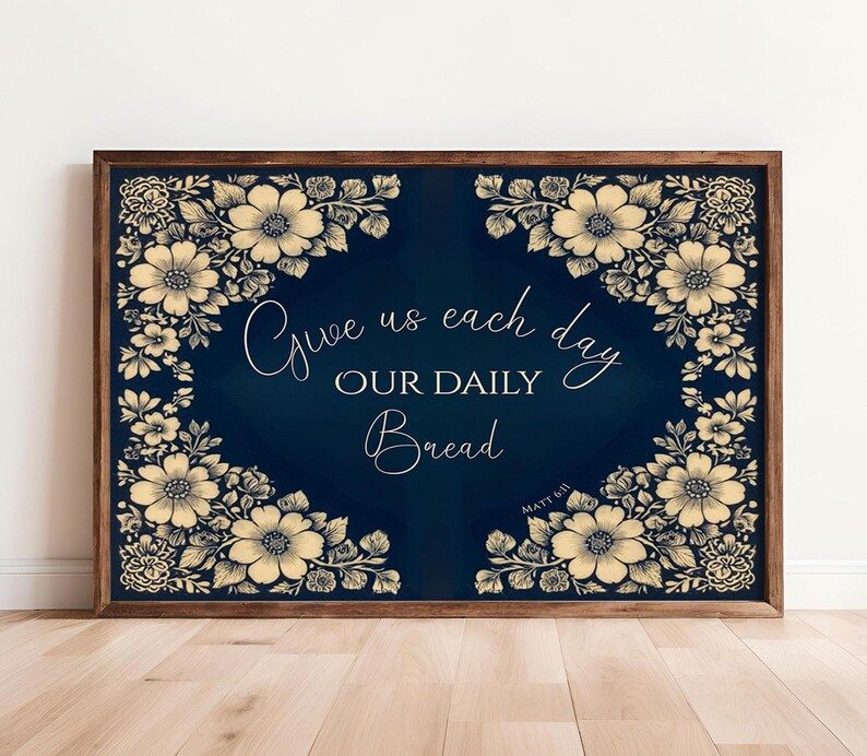 Op de afbeelding: Een ingelijst kunstwerk met een donkerblauwe achtergrond en bloemmotieven in de hoeken. De tekst "Give us each day OUR DAILY Bread" is geschreven in een elegant lettertype. De lijst is donkerbruin.