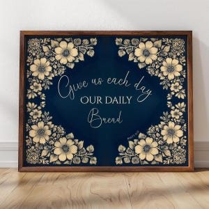 Op de afbeelding: Een ingelijste print met een donkerblauwe achtergrond en bloemenrand. De tekst "Give us each day OUR DAILY Bread" is geschreven in elegant schrift. De lijst is donkerbruin.