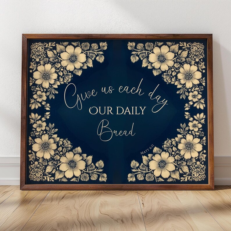 Op de afbeelding: Een ingelijste print met een donkerblauwe achtergrond en bloemenrand. De tekst "Give us each day OUR DAILY Bread" is geschreven in elegant schrift. De lijst is donkerbruin.
