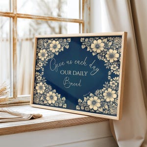 Op de afbeelding: Een ingelijste print met de tekst "Give us each day our daily bread" omringd door bloemenillustraties. Het kunstwerk heeft een marineblauwe achtergrond met cr&egrave;mekleurige bloemen en tekst, in een licht houten frame.