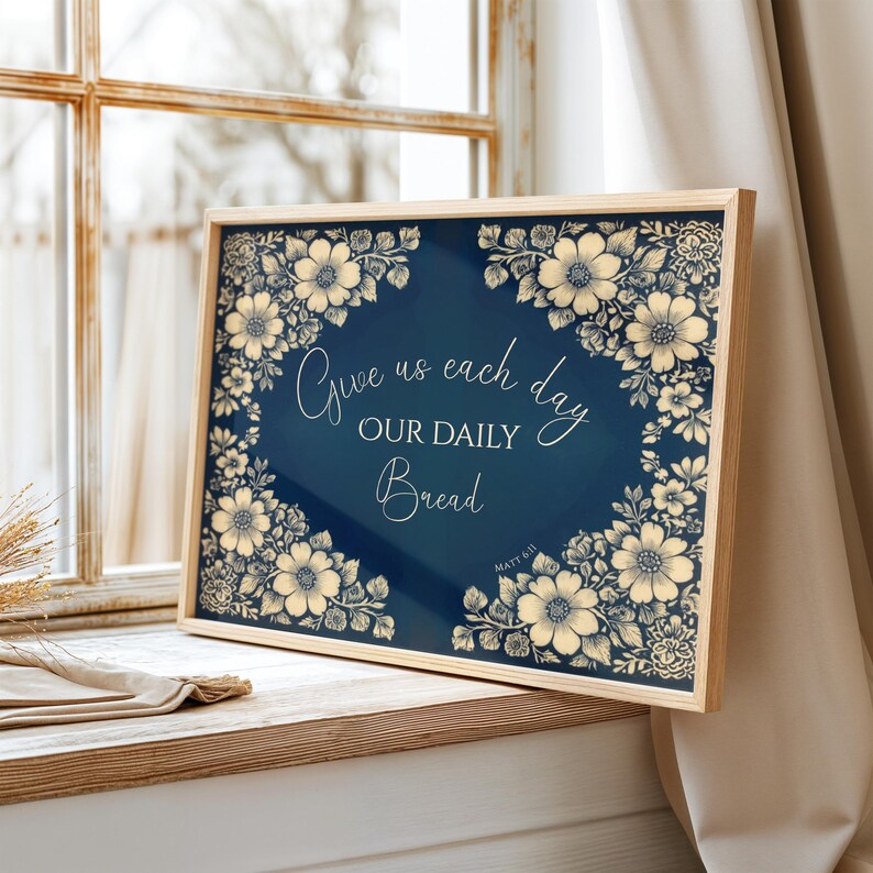Op de afbeelding: Een ingelijste print met de tekst "Give us each day our daily bread" omringd door bloemenillustraties. Het kunstwerk heeft een marineblauwe achtergrond met cr&egrave;mekleurige bloemen en tekst, in een licht houten frame.