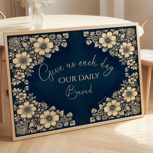 Op de afbeelding: Een ingelijste print met een marineblauwe achtergrond en bloemmotief. De tekst "Give us each day OUR DAILY Bread" staat in sierlijke letters. De bloemenrand is in cr&egrave;me- en beigetinten.