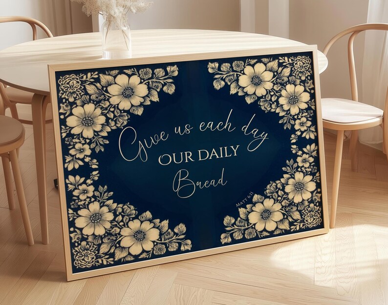 Op de afbeelding: Een ingelijste print met een marineblauwe achtergrond en bloemmotief. De tekst "Give us each day OUR DAILY Bread" staat in sierlijke letters. De bloemenrand is in cr&egrave;me- en beigetinten.