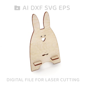 May include: A wooden phone stand shaped like a bunny rabbit. The stand has a cutout for a phone and two small feet. The bunny has a simple face with two eyes and a mouth.