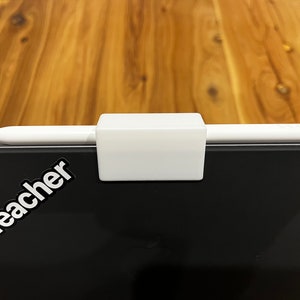 Peut inclure: Un stylet blanc est maintenu par un support rectangulaire blanc. Le support est fixé à une surface noire avec le texte "AppleTeacher" en blanc. L'arrière-plan est une surface en bois.