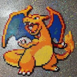 May include: A pixel art depiction of Charizard, a popular character from the Pokémon franchise. The artwork is created using colorful plastic beads, with orange, yellow, blue, and red hues. The wings are blue with a yellow outline, and the body is primarily orange.