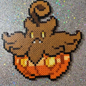 May include: A brown and orange pixel art design of a ghost wearing a pumpkin hat. The ghost has a round head with two eyes and a small mouth. The pumpkin hat has a stem on top and a face with a wide smile and two eyes.