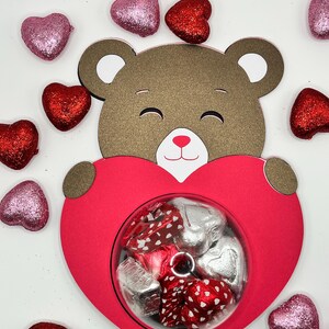 Puede incluir: Un oso de peluche marr&oacute;n sosteniendo una caja roja en forma de coraz&oacute;n llena de caramelos de chocolate. La caja tiene una ventana transparente para ver los caramelos en el interior. El fondo es blanco con corazones de purpurina rosa y rojo esparcidos.