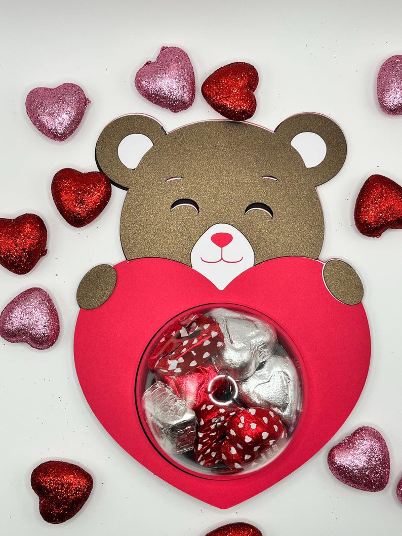 Puede incluir: Un oso de peluche marr&oacute;n sosteniendo una caja roja en forma de coraz&oacute;n llena de caramelos de chocolate. La caja tiene una ventana transparente para ver los caramelos en el interior. El fondo es blanco con corazones de purpurina rosa y rojo esparcidos.