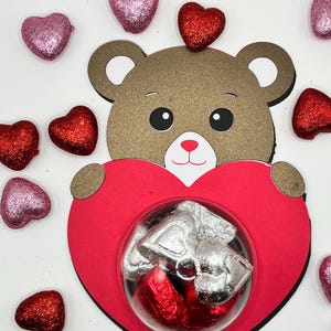 Puede incluir: Un oso de peluche marr&oacute;n sosteniendo una caja roja en forma de coraz&oacute;n con una ventana de pl&aacute;stico transparente. La caja contiene peque&ntilde;os caramelos de chocolate envueltos en papel de aluminio plateado. El fondo es blanco con corazones de purpurina rojos y rosas esparcidos.