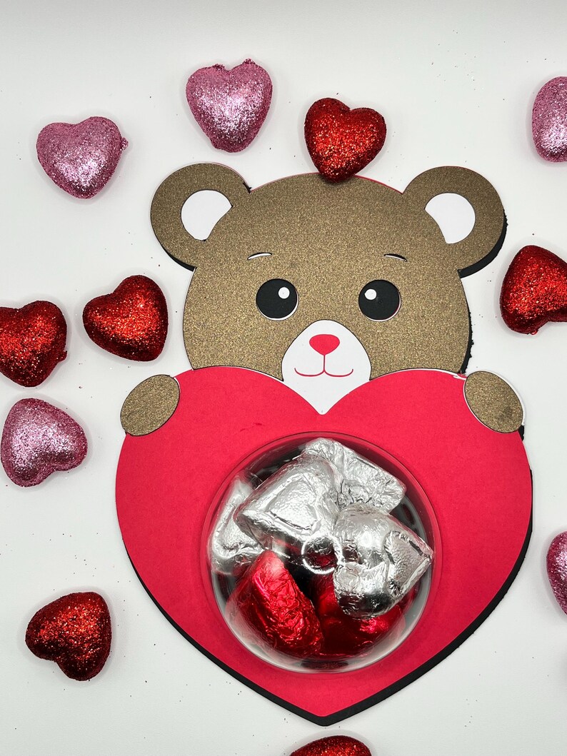 Puede incluir: Un oso de peluche marr&oacute;n sosteniendo una caja roja en forma de coraz&oacute;n con una ventana de pl&aacute;stico transparente. La caja contiene peque&ntilde;os caramelos de chocolate envueltos en papel de aluminio plateado. El fondo es blanco con corazones de purpurina rojos y rosas esparcidos.