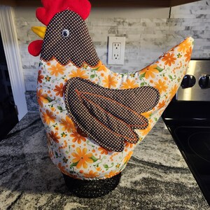 May include: A fabric chicken-shaped tea cozy featuring a white body with an orange floral pattern, a brown and white polka-dot head and wing, a bright red felt comb and wattle, a yellow beak, and a single googly eye attached to the side of the head.