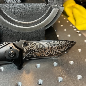 May include: A black folding knife with an ornate, engraved floral pattern on the blade. The knife is open, revealing the detailed design. The handle and blade are dark, contrasting with the lighter engraving. The knife is on a metal surface.