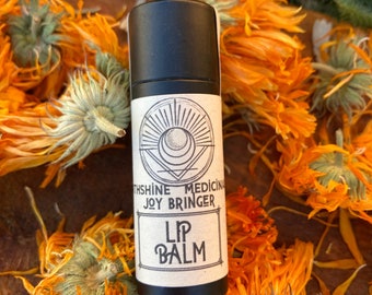 May include: A black tube of lip balm with a white label that says "Moonshine Medicina Joy Bringer Lip Balm". The label has a sun and moon design. The tube is surrounded by dried orange flowers.