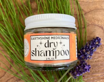 May include: A small glass jar of Earthshine Medicinals dry shampoo with a white screw-top lid. The orange label features black text reading Earthshine Medicinals, dry shampoo, and 2 fl oz, accented by small star symbols.