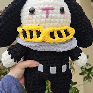 May include: A handmade crochet plushie in the shape of a bunny. The bunny has a white head, black ears, and a black body. It wears a yellow and gray scarf and has black eyes and a pink nose.
