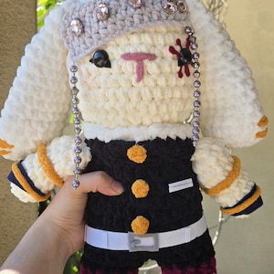 May include: A handmade crochet plush toy in the shape of a bunny. The bunny has white fur, a gray head covering with rhinestones, and a black and orange outfit. The bunny has a pink nose and black eyes.
