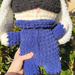 May include: A crocheted plush toy rabbit wearing a blue jumpsuit and a black eye mask. The rabbit has white fur and long floppy ears.