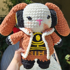 May include: A crocheted plush toy in the shape of a bunny wearing a brown jacket with yellow and black accents. The bunny has white fur, brown ears, and black eyes.