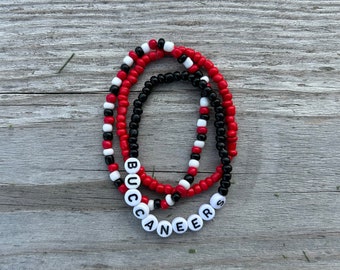 Puede incluir: Dos pulseras de cuentas con cuentas negras, rojas y blancas. Las pulseras deletrean "BUCCANEERS" en letras blancas.