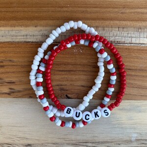 May include: Three beaded bracelets in red, white, and gray with the word "BUCKS" spelled out in white beads.