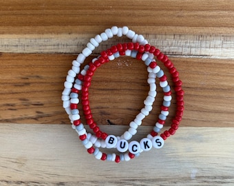 May include: Three beaded bracelets in red, white, and gray with the word "BUCKS" spelled out in white beads.