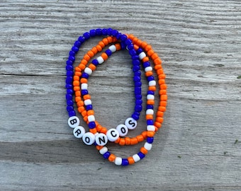 May include: Three beaded bracelets in orange, white, and blue with the word "BRONCOS" spelled out in white beads.