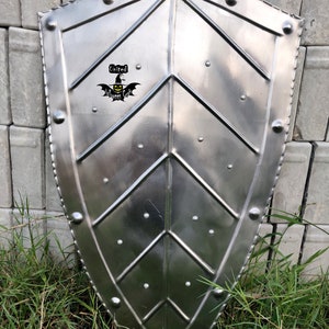 May include: A silver metal shield with a pointed bottom and a decorative design of overlapping triangles. The shield has a black and yellow sticker with the text "United Halloween Craft" on it.