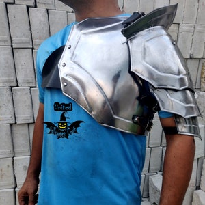 May include: A silver metal shoulder armour piece with a black leather strap. The armour is worn by a person wearing a blue t-shirt with the text "United Halloween Craft".