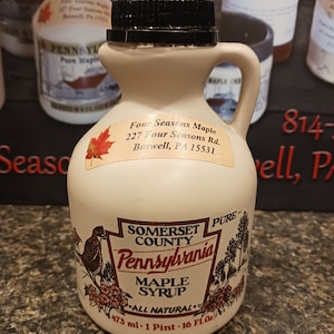 May include: A white plastic bottle of Somerset County Pennsylvania pure maple syrup. The bottle is labeled with the text "Somerset County Pennsylvania Pure Maple Syrup" and "All Natural". The bottle is 16 fluid ounces or 473 milliliters.