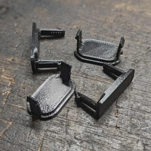 Può includere: Quattro componenti in plastica nera stampati in 3D, ciascuno caratterizzato da una base rettangolare arrotondata piatta con una linguetta verticale perpendicolare che si estende da un lato. Le superfici mostrano una texture a strati.