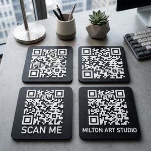 Può includere: Quattro sottobicchieri quadrati neri con codici QR bianchi in rilievo. Due sottobicchieri mostrano solo il codice QR, mentre quello in basso a sinistra include il testo SCAN ME e quello in basso a destra il testo MILTON ART STUDIO in bianco.