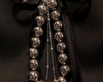 May include: A silver chain with a series of large, round, silver beads. The chain is attached to a black leather bag with a black ribbon.