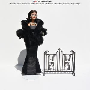 May include: A doll in a black gown and fur wrap, with a pearl necklace and earrings. A black metal decorative bed frame is next to the doll. The doll is on a white stand.
