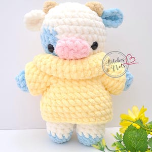 May include: A handmade plush cow with a white, blue, and pink face, wearing a yellow sweater. The cow has black eyes and small brown ears. The text "Stitches & Nots" is visible on the right side of the image. The plush toy is on a white background.